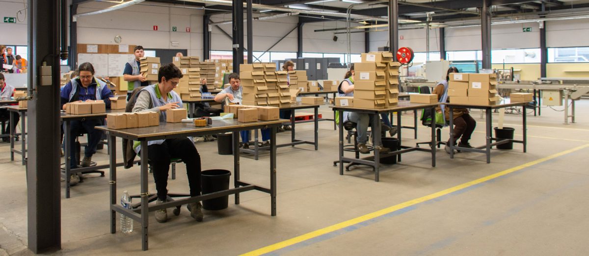 Medewerkers van Bewel Sint-Truiden zitten aan inpaktafels en verpakken er producten in kleine dozen.