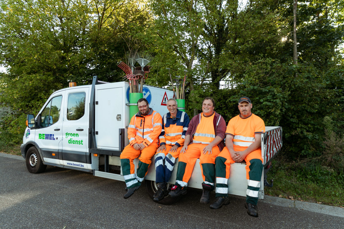Een team van de groendienst van Bewel Diepenbeek zit in de laadbak van een Bewel camionette. Ze zijn met vier en lachen naar de camera.