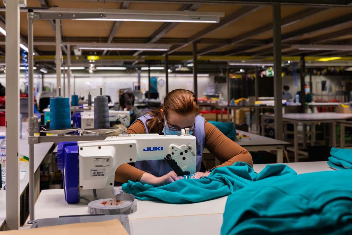 Een medewerker van Bewel Hasselt zit achter een naaimachine, in de stikkerij. Ze is aan het werk met een felblauwe fleece.