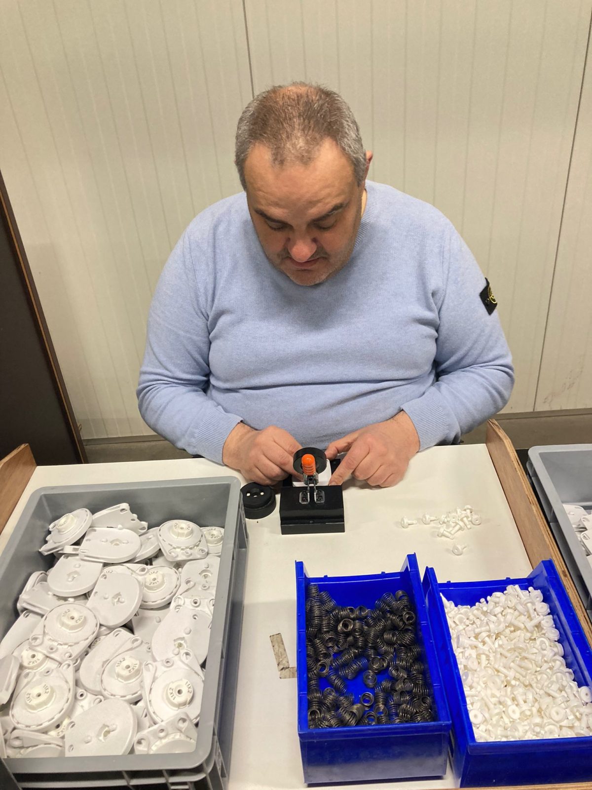 Een medewerker van Bewel assembleert kleine onderdeeltjes voor Hunter Douglas. Voor hem staan bakken met verschillende onderdeeltjes om jaloezieën te assembleren..