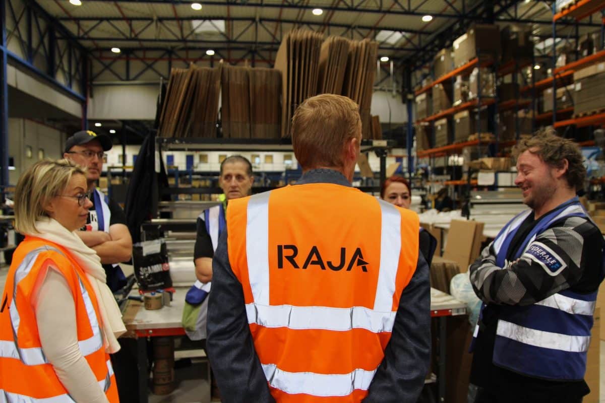 De medewerkers van Bewel en RAJA staan in het magazijn van RAJA te praten. Centraal staat een man met zijn rug naar de camera. Op de achterkant van zijn fluo-oranje hesje staat in het groot 'RAJA' te lezen.